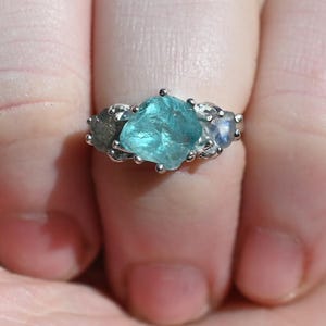 May include: A silver ring with a large, irregular, light blue gemstone at its centre, accompanied by two smaller, round, light blue stones. The ring is displayed on a hand.