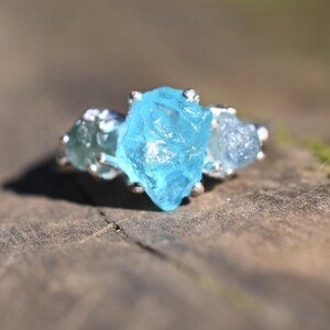 Pode incluir: Um anel de prata com uma grande pedra preciosa azul clara no centro, ladeada por duas pedras menores e mais claras. O anel está sobre uma superfície de madeira desfocada, com a luz do sol criando um brilho suave. É uma joia.