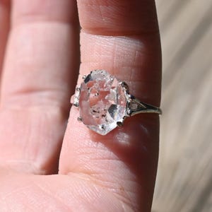 May include: A clear, faceted gemstone ring with a silver band. The gemstone is set in a prong setting and has a unique, irregular shape. The ring is displayed on a person's finger.