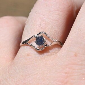 Pode incluir: Um anel de tom prateado com uma pequena pedra preciosa azul-escura de corte bruto engastada no centro. A banda tem um design delicado, ondulado e aberto que emoldura a pedra, criando uma silhueta distinta com um acabamento metálico.