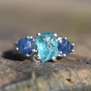 Pode incluir: Um anel de prata com três pedras brutas. A pedra central é azul claro, ladeada por duas pedras azuis mais escuras. O anel está sobre uma superfície de madeira envelhecida, com foco na joia.