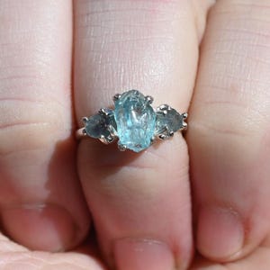 May include: A silver ring with three pale blue gemstones. The central stone is an elongated oval, with two smaller, triangular stones either side. The ring is shown on a person's hand.