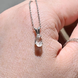Pode incluir: Um colar de prata com um pingente transparente em forma de gota suspenso por uma corrente delicada. O pingente é mantido em uma montagem de metal simples e uma corrente de prata fina correspondente está enrolada ao redor do dedo.