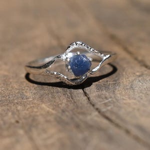 May include: A silver ring featuring a textured, organic band that splits to hold a single, small, raw blue gemstone at its center. The metal surface of the band has a rough, uneven finish, contrasting with the smooth, rounded shape of the blue stone.