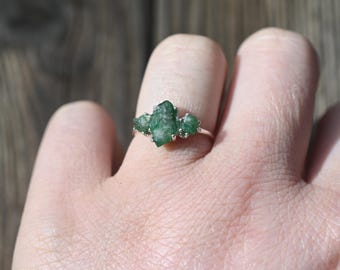 May include: A silver ring featuring three raw emerald stones set in a cluster. The ring is worn on a finger.