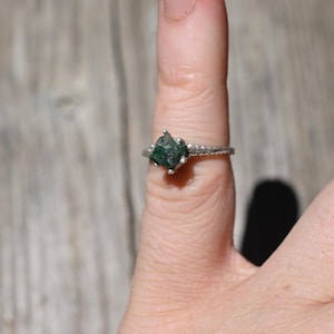 Pode incluir: Um anel de prata com uma pedra preciosa verde bruta e não cortada. A banda do anel tem um design texturizado. A pedra preciosa é o ponto focal, mostrando sua forma natural irregular e sua cor verde escura. O anel é exibido em um dedo.