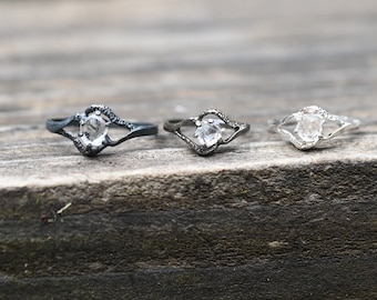 May include: Three silver rings with a clear gemstone set in a delicate, open design. The rings are on a wooden surface.
