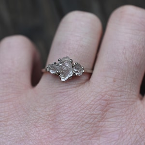 May include: A silver ring featuring three clear, irregularly shaped gemstones clustered together. The ring is set on a slender band and is displayed on a hand, showcasing the jewelry's design and details. The gemstones have a raw, natural appearance.