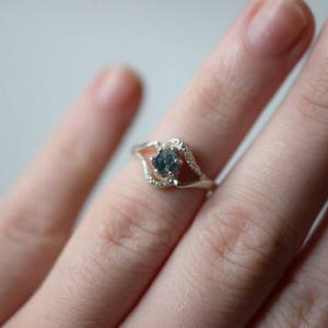 May include: A silver ring with a delicate, open design features a round, gray gemstone in the center. The ring is worn on a finger.