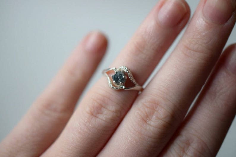 May include: A silver ring with a delicate, open design features a round, gray gemstone in the center. The ring is worn on a finger.