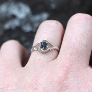May include: A silver ring with a dark blue gemstone. The ring has a delicate, openwork design around the stone, with small details. The ring is on a finger, with a blurred background.