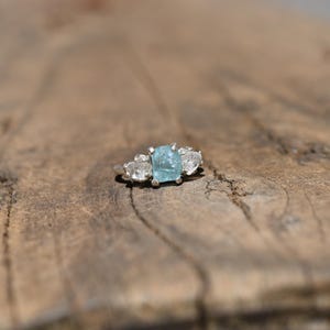 Pode incluir: Um anel de prata com uma pedra preciosa central de corte bruto em azul claro, ladeada por duas pedras mais pequenas, transparentes e em forma de pera. O anel apresenta um engaste de garras e uma banda de metal polido.