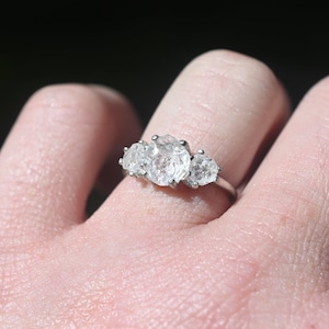 May include: A silver ring with three clear oval-shaped gemstones set in a row. The ring is on a person's finger.