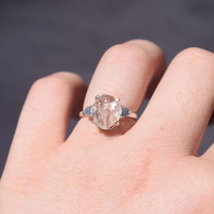 May include: A silver ring with a large, clear, irregularly shaped central gemstone and two smaller blue gemstones. The ring is displayed against a plain background.