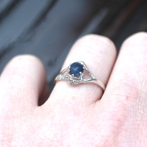 May include: A silver ring featuring a dark blue gemstone. The ring has a detailed, nature-inspired design with small accent stones. The ring is displayed on a finger, showcasing its elegant and intricate craftsmanship.