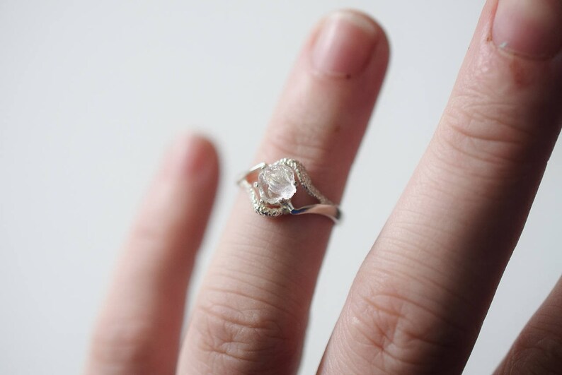 May include: A silver ring with a clear, faceted, raw crystal stone set in the center. The ring has a delicate, curved design.