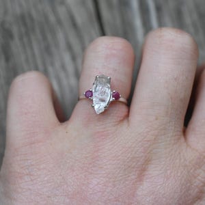 Op de afbeelding: Een zilveren ring met een heldere, gefacetteerde, langwerpige edelsteen en twee kleinere, ronde, roze edelstenen aan weerszijden.