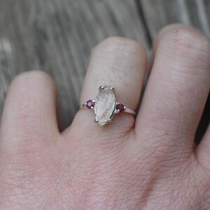 Op de afbeelding: Een zilveren ring met een heldere, ruwe kristalsteen en twee kleine robijnstenen aan weerszijden.