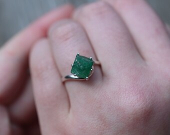 May include: A silver ring featuring a vibrant green emerald-cut gemstone. The ring's band curves gracefully, and the emerald is held in place by four prongs. The gemstone's color is a rich, deep green.