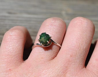 Puede incluir: Un anillo de plata con una esmeralda en bruto. La esmeralda es de color verde oscuro y está sujeta por delicadas garras. La banda del anillo tiene un diseño sencillo y elegante.