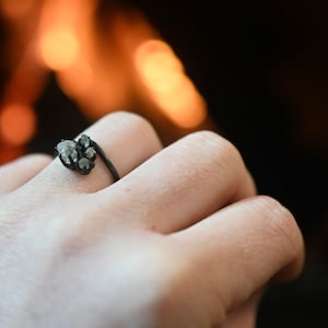 May include: A dark-toned metal ring featuring a cluster of small, raw, translucent, light-colored stones set in a bypass style. The band is thin and dark, contrasting with the pale, irregular shapes of the stones held in a dark metal setting.