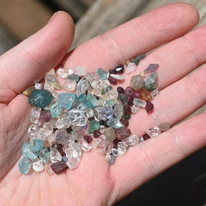 May include: A handful of small, raw gemstones in various colors, including clear, blue, green, purple, and red. The gemstones are irregularly shaped and appear to be uncut. The image is taken in natural light, highlighting the gems' clarity.