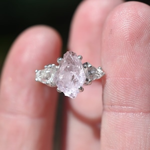 May include: A silver ring with a large, clear, pink crystal set in the center. Two smaller, clear crystals are set on either side of the center crystal.