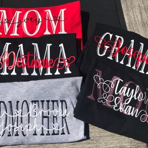 May include: Four t-shirts with different titles and names. The first shirt is red with white text that says "MOM" with the name "Joey" in a heart. The second shirt is black with white text that says "MAMA" with the name "Destanie" in a heart. The third shirt is black with white text that says "GRAMMY" with the name "Joseph" in a heart. The fourth shirt is gray with white text that says "GODMOTHER" with the names "Evan, Brook, Joseph" in hearts.