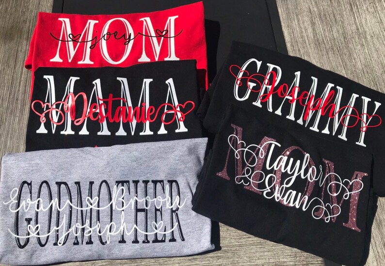 May include: Four t-shirts with different titles and names. The first shirt is red with white text that says "MOM" with the name "Joey" in a heart. The second shirt is black with white text that says "MAMA" with the name "Destanie" in a heart. The third shirt is black with white text that says "GRAMMY" with the name "Joseph" in a heart. The fourth shirt is gray with white text that says "GODMOTHER" with the names "Evan, Brook, Joseph" in hearts.