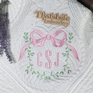 May include: White quilted blanket with a pink bow and green leaf embroidery. The initials "CSJ" are embroidered in pink within a green leaf wreath. The text "Thatslife Embroidery" is visible.