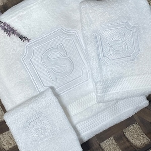 May include: Three white towels with embroidered letter 'S' in a decorative frame. The towels are folded and arranged in a wooden basket.