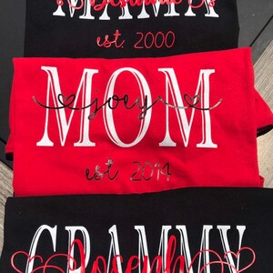 May include: Three black t-shirts with white text and red hearts. The top shirt says "MAMA Destanie est. 2000". The middle shirt says "MOM joey est. 2014". The bottom shirt says "GRAMMY Joseph est. 2014".