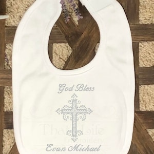 Puede incluir: Babero blanco para beb&eacute; con una cruz bordada en plata y el texto "God Bless" y "Evan Michael".