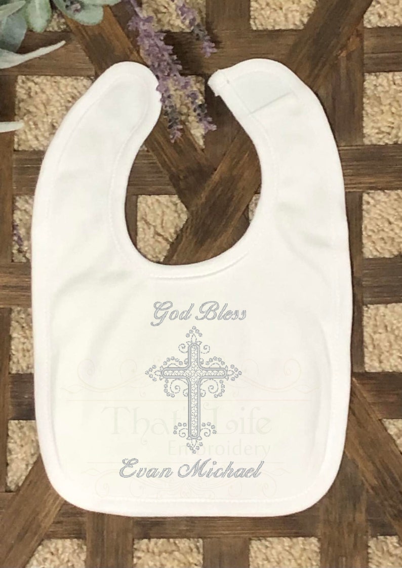 Puede incluir: Babero blanco para beb&eacute; con una cruz bordada en plata y el texto "God Bless" y "Evan Michael".