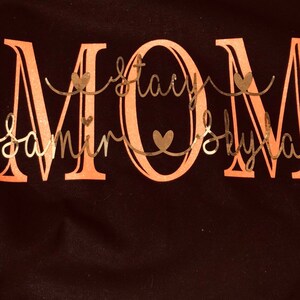 May include: A black t-shirt with the word "MOM" in large, pink letters. The names "Samir", "Stacy", and "Skylan" are written in cursive script below the word "MOM" in a gold color.