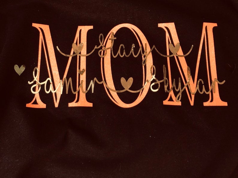 May include: A black t-shirt with the word "MOM" in large, pink letters. The names "Samir", "Stacy", and "Skylan" are written in cursive script below the word "MOM" in a gold color.