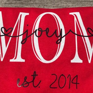 May include: Red t-shirt with white glitter lettering that says "MOM" with the word "joey" written in cursive between the "M"s.  The shirt also has black lettering that says "est. 2014" below the word "MOM".