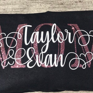 May include: A black t-shirt with a pink glitter MOM design. The names "Taylor & Evan" are written in white cursive lettering.