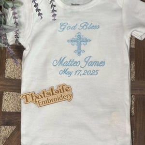Puede incluir: Mono blanco para beb&eacute; con bordado azul de una cruz, el texto "God Bless Matteo James May 17, 2025" y una etiqueta de madera que dice "Thatslife Embroidery".