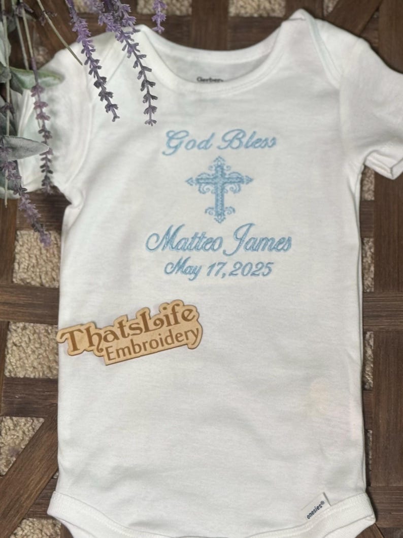 Puede incluir: Mono blanco para beb&eacute; con bordado azul de una cruz, el texto "God Bless Matteo James May 17, 2025" y una etiqueta de madera que dice "Thatslife Embroidery".