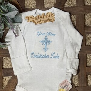 Puede incluir: Mono blanco de manga larga para beb&eacute; con una cruz bordada en azul y el texto "God Bless Christopher Luke".