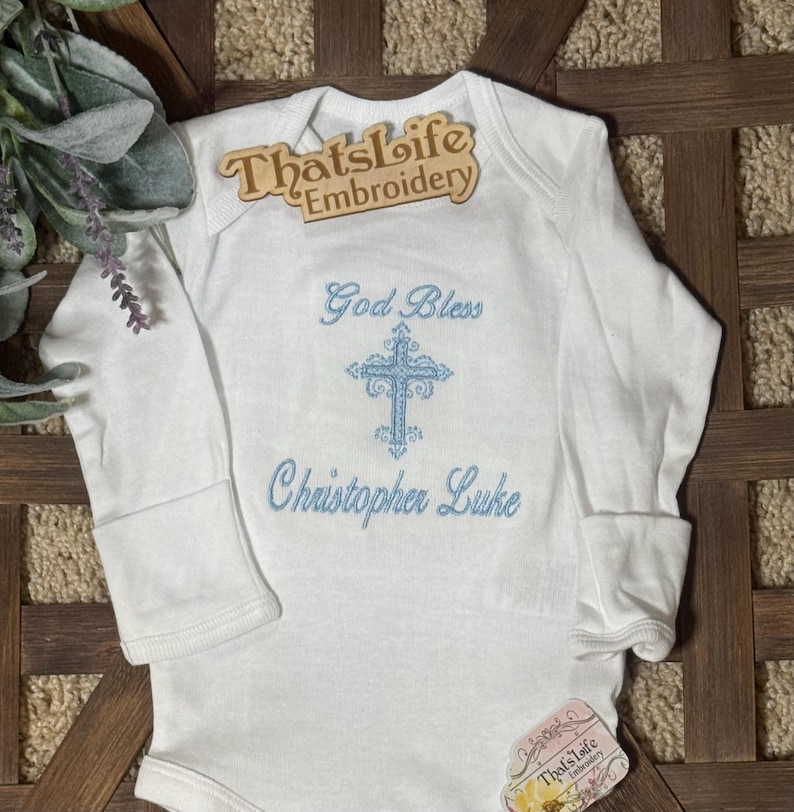 Puede incluir: Mono blanco de manga larga para beb&eacute; con una cruz bordada en azul y el texto "God Bless Christopher Luke".