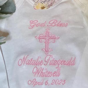 Puede incluir: Un body blanco para beb&eacute; con bordado rosa que dice "God Bless Natalie Fitzgerald Whitesell April 6, 2025" y una cruz rosa.