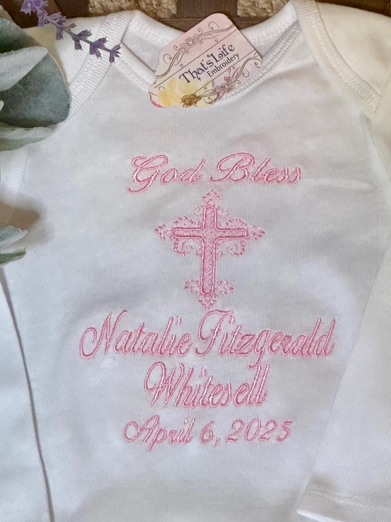 Puede incluir: Un body blanco para beb&eacute; con bordado rosa que dice "God Bless Natalie Fitzgerald Whitesell April 6, 2025" y una cruz rosa.