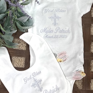 Puede incluir: Mono y babero blancos para beb&eacute; con bordado plateado. El bordado incluye una cruz, las palabras "God Bless" y el nombre "Myles Patrick" con la fecha "March 22, 2025".