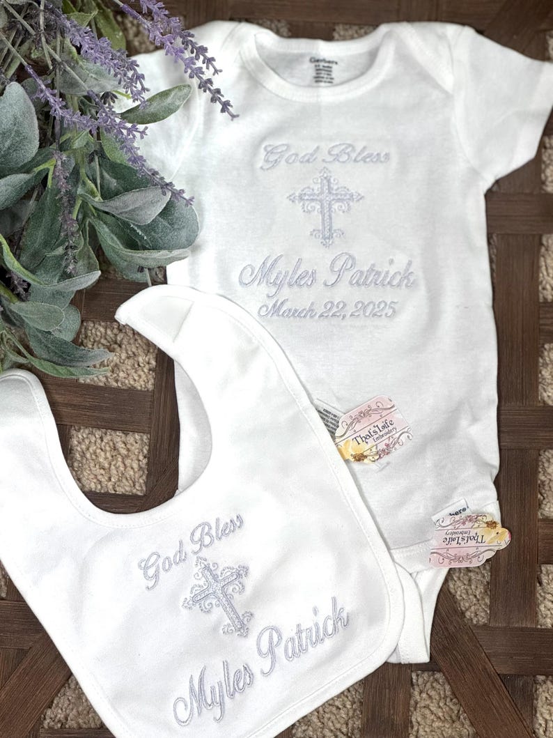 Puede incluir: Mono y babero blancos para beb&eacute; con bordado plateado. El bordado incluye una cruz, las palabras "God Bless" y el nombre "Myles Patrick" con la fecha "March 22, 2025".