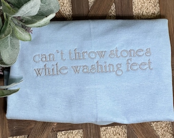 May include: Light blue t-shirt with the text "can't throw stones while washing feet" embroidered in silver thread.