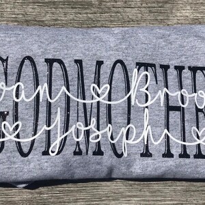 May include: Gray t-shirt with the text "GODMOTHER" in black with white cursive writing underneath that says "Evan Brooke Joseph" with three white hearts.