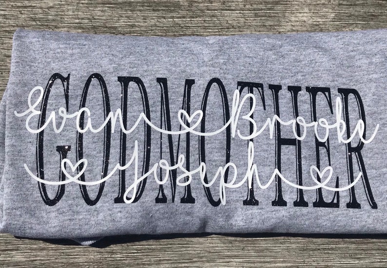May include: Gray t-shirt with the text "GODMOTHER" in black with white cursive writing underneath that says "Evan Brooke Joseph" with three white hearts.
