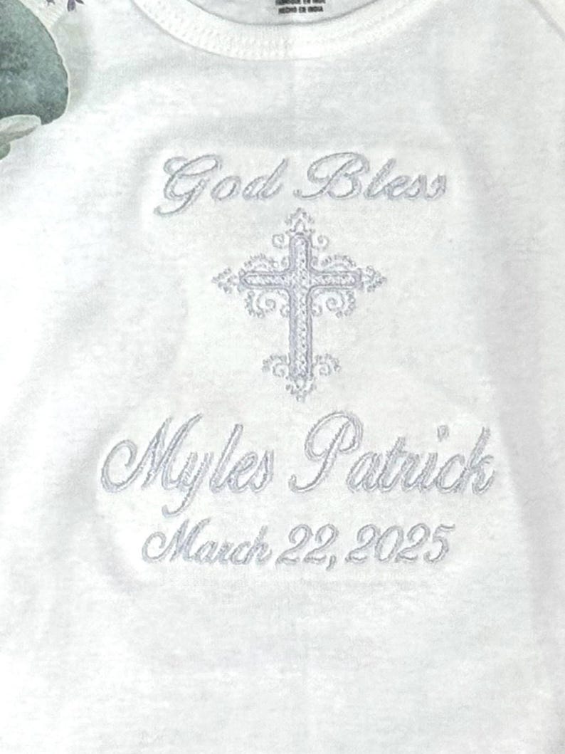 Puede incluir: Unisie blanco para beb&eacute; con bordado plateado. El bordado dice "God Bless Myles Patrick March 22, 2025" e incluye una cruz.